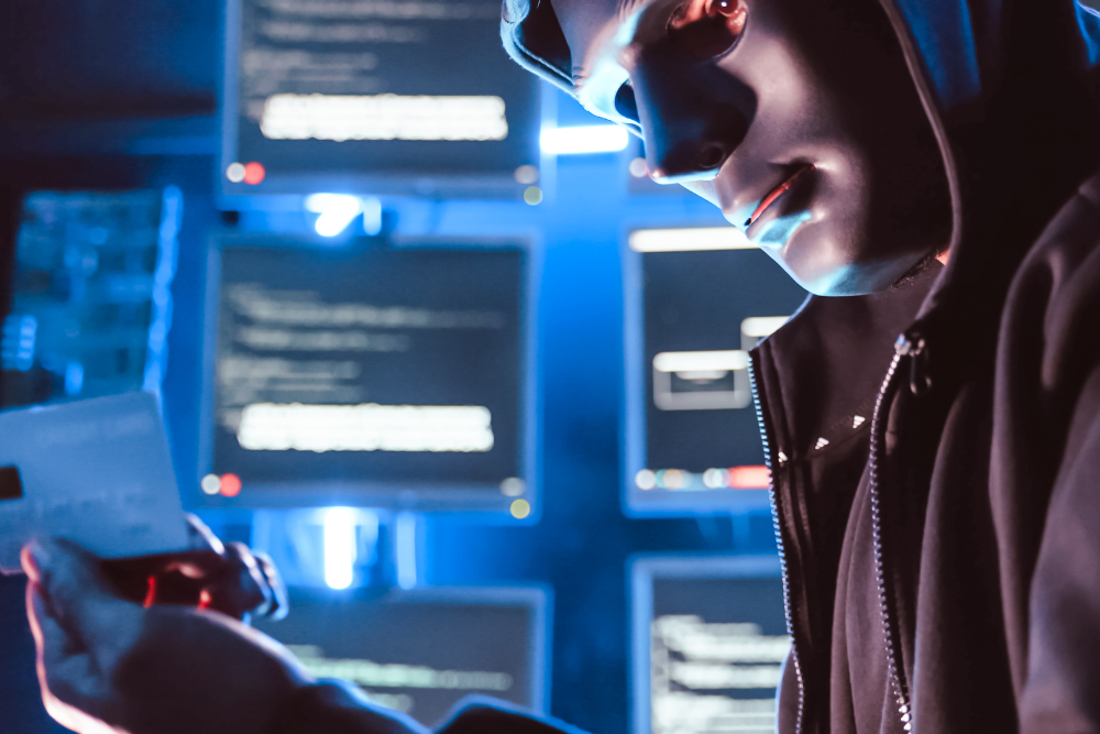 Masked hacker holding a credit card by code screens, showing phishing and data theft and how to prevent cyberattacks.